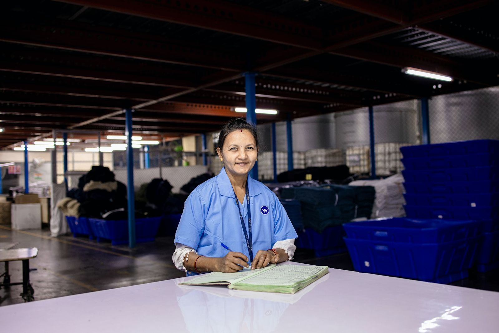 Indian woman in a blue work coat smiling at a table on a factory floor, a notebook in front of her