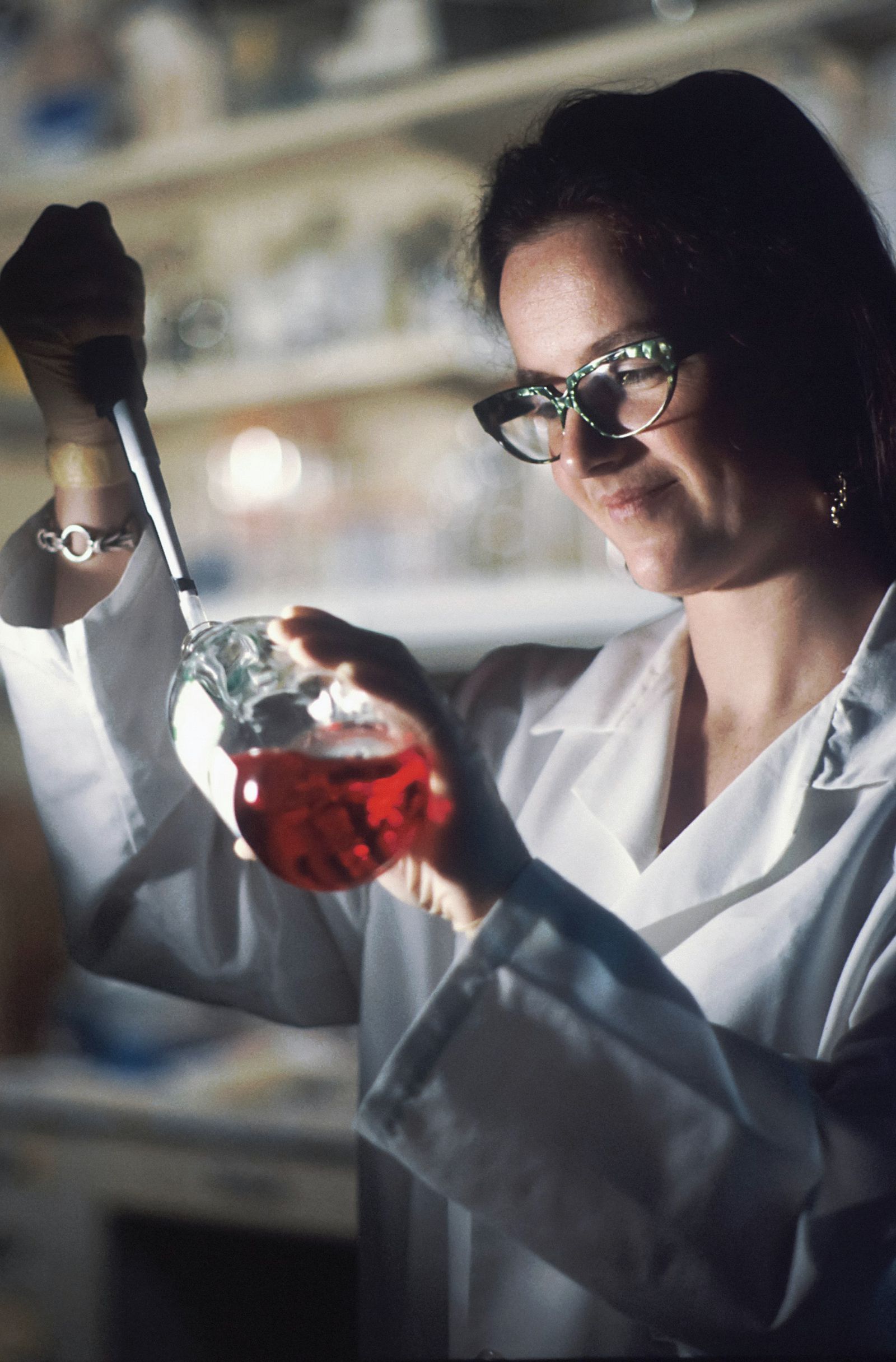 Woman scientist in a lab coat and glasses holding a flask of red culture medium in a laboratory